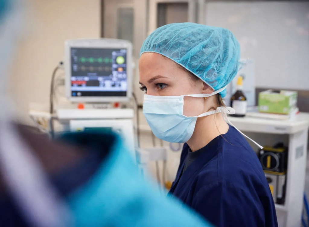 CRNA in a surgical cap and mask standing beside an anesthesia monitor in the operating room